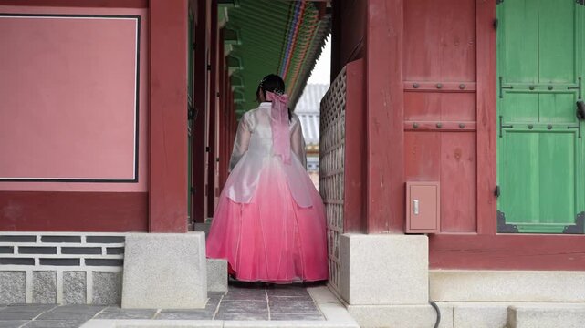 Woman in flowing pink hanbok steps through traditional gate. Her gaze ahead suggests calm contemplation. Soft light highlights the vibrant color of her garment