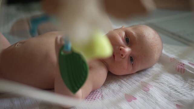 Adorable newborn baby with blue eyes lying in a cozy crib and interacting with a colorful hanging mobile, the curious infant touching the toy while developing motor skills and exploring the world