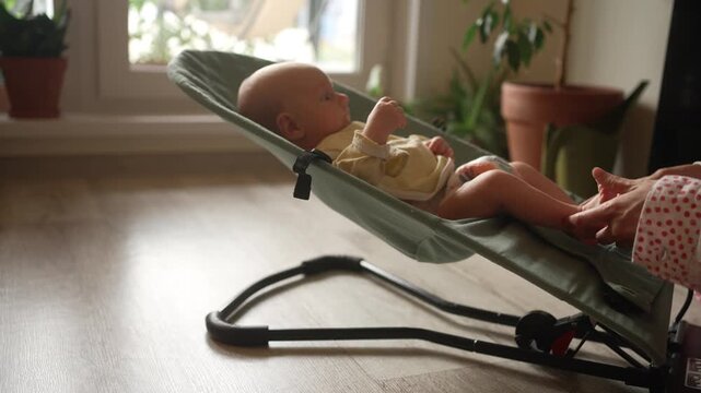 Mother's hands gently massaging her newborn infant's tiny feet while the baby rests in a comfortable bouncer chair, a tender bonding activity promoting sensory stimulation in a home setting