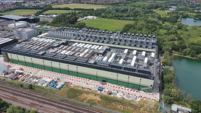 An expansive data center building is observed from an aerial viewpoint. It stands next to active railway lines, a large body of water, and surrounding green landscapes.
