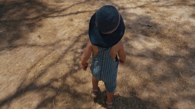 Top-down full shot of a toddler in a wide sun hat and striped romper standing on parched straw-covered ground. Conveys childhood, heat and rural summer life.