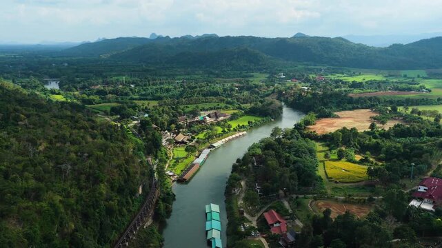 Aerial ascend overview of Krasae River and railway winding across lush valley in Kanchanaburi Thailand, drone