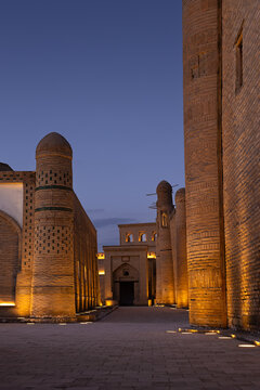 Ichan Kala old town in Khiva illuminated at twilight with brick minarets and ornamental walls. Empty passage between historic buildings creates a dramatic perspective under a deep blue sky.