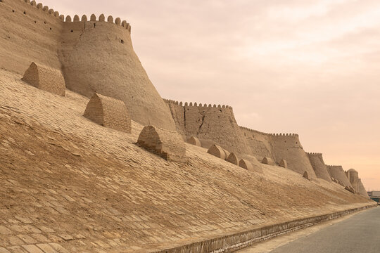 Massive fortress walls of Ichan Kala in Khiva with crenellated towers and mud-brick buttresses at sunset. Ideal for travel publications, history textbooks, and cultural heritage materials.