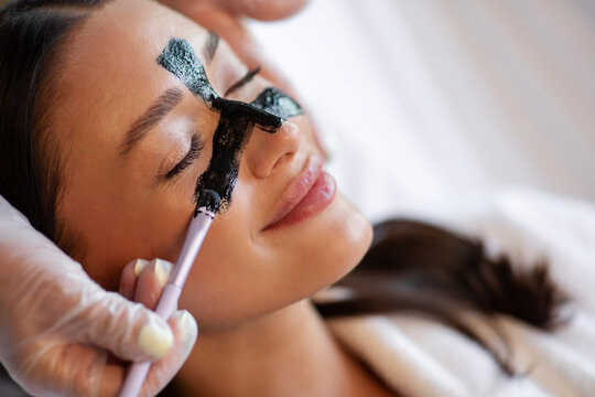 Closeup of woman enjoying professional skincare session while beauty specialist applying purifying charcoal mask to T-zone for deep cleansing