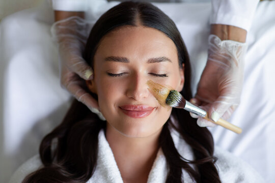 Smiling woman enjoying calming facial beauty procedure while specialist applying skincare mask using soft cosmetic brush, top view. Holistic skin wellness