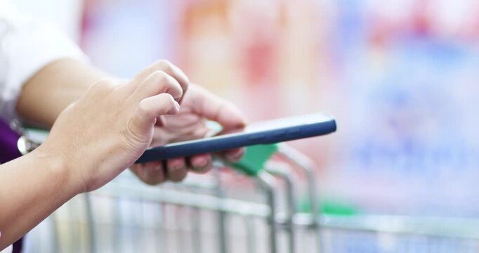 Customer using a smartphone while shopping in a supermarket with a shopping cart. Mobile shopping, retail, price comparison, digital payment, and modern lifestyle