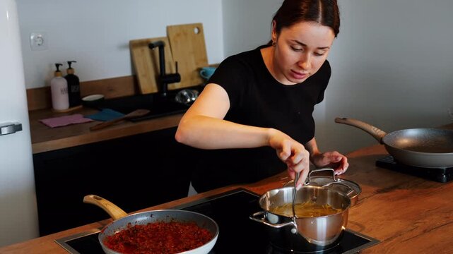 A woman stirs boiling pasta in a pot while bolognese sauce simmers in a pan beside it on an induction hob in a modern home kitchen.