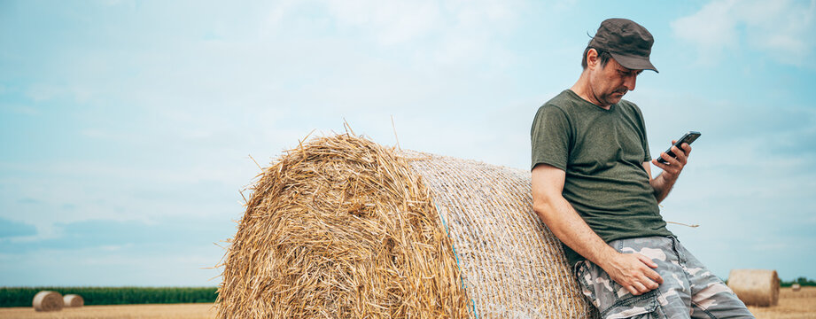 Farmer using a smartphone while leaning against a large round hay bale in a harvested field. Smart farming, digital agriculture and post-harvest farm management concept.