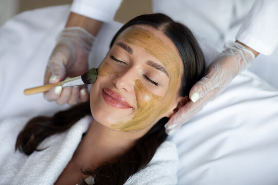 Happy woman receiving professional facial mask as beauty specialist gently treating her skin, closeup shot