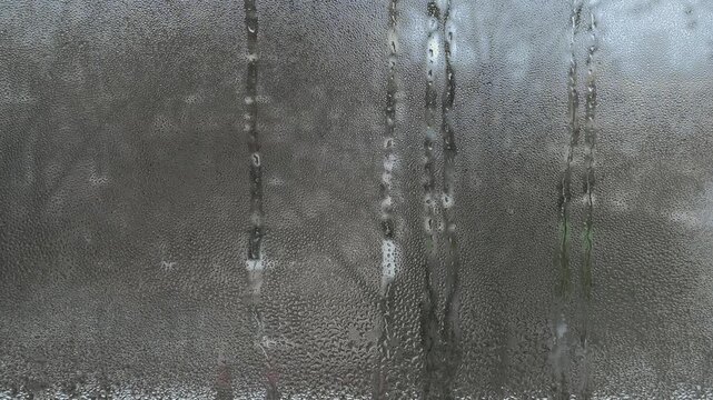Foggy window glass with condensation forming water droplets and streaks. Moist glass surface reflects cold, rainy weather outside, creating a misty, blurred view. Indoor atmosphere, wet glass detail,