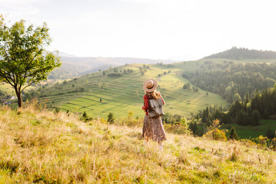 Woman traveler, wearing hat and backpack, sits in sunny green meadow. Beautiful woman in hat feels free and enjoys mountain landscape at sunset. Concepts of relaxation, travel, and enjoyment.