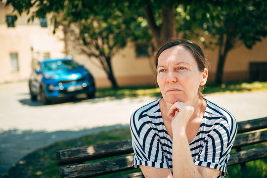 Middle-aged woman sitting on a park bench with a slightly worried expression. Concern, uncertainty and everyday life challenges in an urban environment.