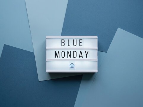 Illuminated lightbox displaying "BLUE MONDAY" with a subtle sad face emoji, set against a textured background of layered cool blue papers