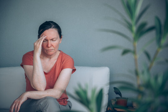 Mature woman suffering from severe headache while sitting on balcony sofa, seen through oleander leaves. Migraine, stress and health problem concept.