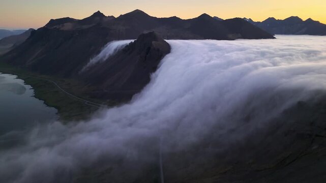 Wide aerial: fog spills down jagged Vestrahorn slopes at golden hour during a rare dawn inversion, East Iceland