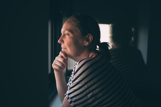 Contemplative middle-aged woman rests her chin on her hand and gazes out the window, captured in soft natural light that emphasizes introspection, solitude, and quiet reflection,