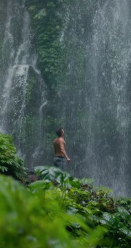 Handsome man with raised arms under powerful waterfall, feeling energy in tropical jungle nature