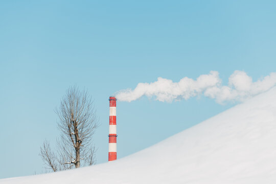 Winter scene of a red-and-white striped factory chimney releasing dense white smoke into clear blue sky over a snow-covered hill, highlighting air pollution from heating industry