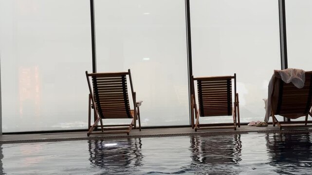 Wooden sun loungers line the spa center pool deck before expansive panoramic windows revealing stunning city skyline views and distant horizon