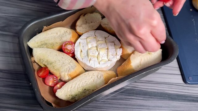 Woman in cozy kitchen attire prepares baked Camembert cheese, stuffing garlic slices into crisscross cuts on the rind, surrounding it with halved tomatoes and baguette chunks for oven roasting.