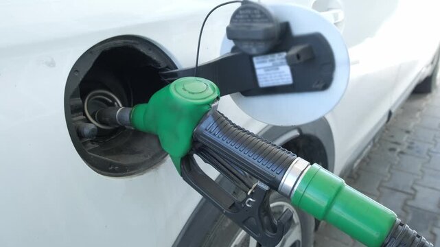 A horizontal close up shot of a green petrol pump nozzle fueling a white crossover vehicle at a gas station, perfect for automotive services, travel logistics, and energy industry marketing.