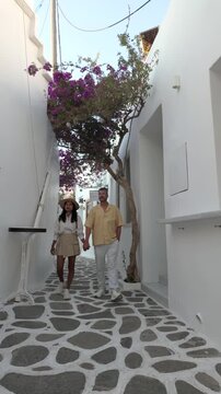 A couple strolls through the beautiful, narrow streets of Naousa, Paros, Greece, where blooming flowers add color to whitewashed buildings. Enjoy the charm of the Cyclades 