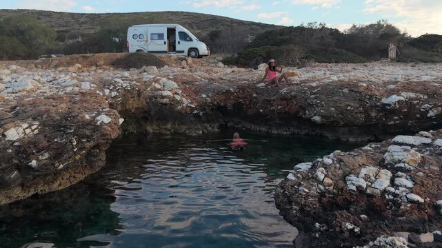 Visitors enjoy the clear waters of Paros Island, diving into a serene swimming spot surrounded by rocky shores. A couple relaxes by the ocean with a camper van motorhome in the background