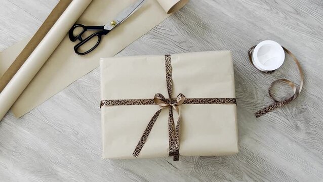 Gift-wrapped package with brown paper and leopard print ribbon, surrounded by scissors, wrapping paper roll, and tape on a wooden surface