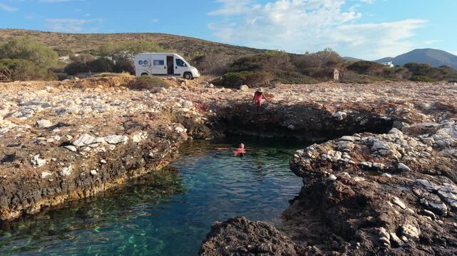 Paros Island, Greece, has sunlit rocky shores and vibrant turquoise waters. Watch as locals dive into the refreshing sea. A couple relaxes by the ocean with a camper van motorhome in the background
