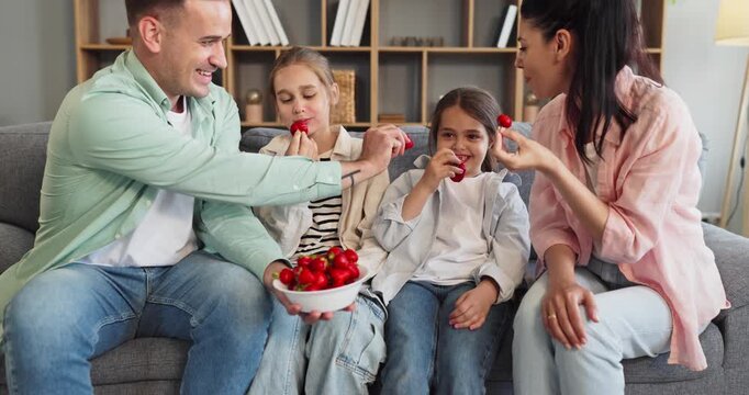 Parents and children eating healthy strawberries together home. Family relaxing on sofa with a bowl of fresh berries smiling and chatting in a cozy living room. Family snack theme for health.
