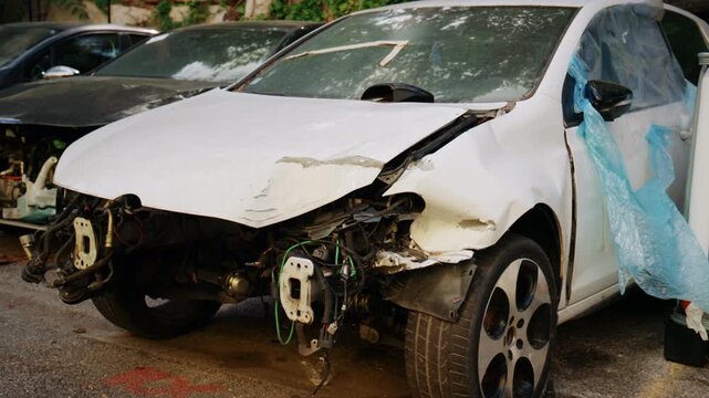 Severely damaged white car with missing front bumper and exposed mechanical parts parked in a scrapyard