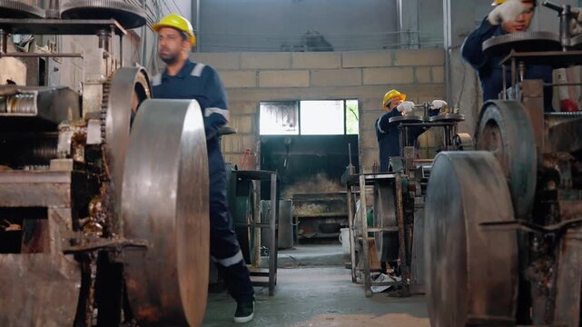 Industrial workers wearing safety helmets operate heavy mechanical equipment inside a manufacturing factory. Teamwork, production process, engineering, and industrial manufacturing concept.
