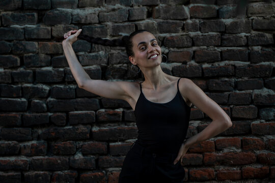 Happy young Hispanic woman holding her long braid, looking at camera and smiling against brick wall.