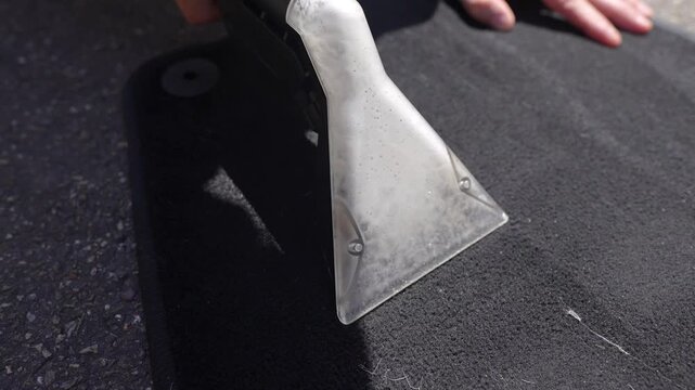 Metal extractor nozzle pulling soapy water from dark rubber car floor mat while sunlight highlights wet texture, cleaning streaks and dirty residue during vehicle detailing