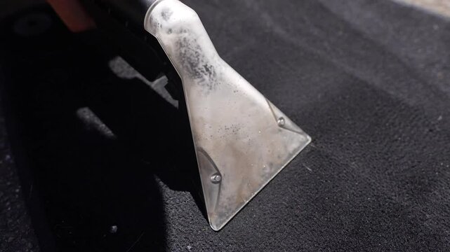 Metal extractor nozzle pulling soapy water from dark rubber car floor mat while sunlight highlights wet texture, cleaning streaks and dirty residue during vehicle detailing