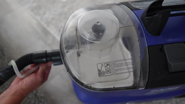 Close oblique top view shows blue canister vacuum cleaner working on carpet while hand presses control near hose connection, mist rises from vent, and clear chamber reveals inner filter Top view of bl