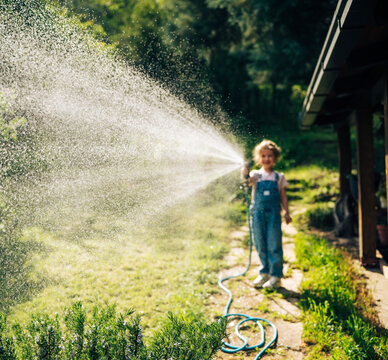Atmospheric out of focus shot of a little 5-year-old girl in overalls watering backyard lawn with hose.