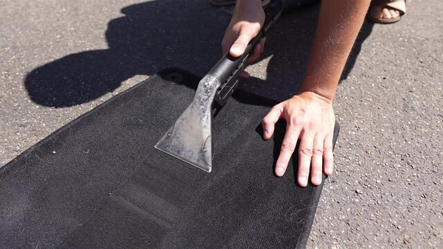 Handheld extractor cleaning dark automotive floor mat on sunlit asphalt, water and foam lifting from fibers while hands steady surface during close-up maintenance demonstration Overhead close-up shows