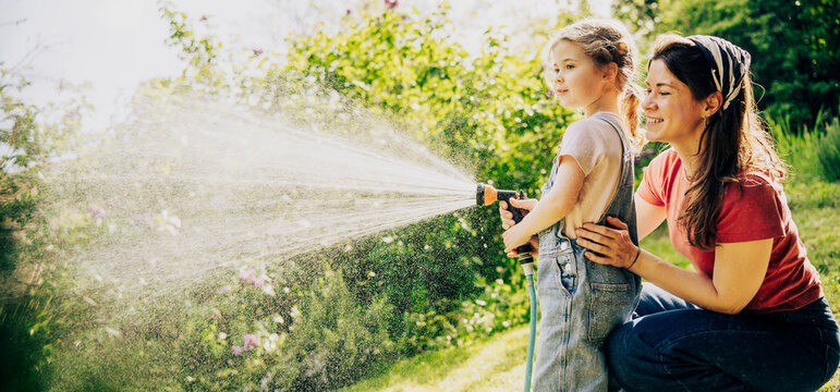 Happy mother and little daughter in denim overalls laughing and watering lawn with hose together.