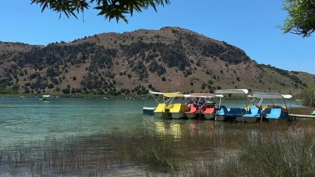 Colorful pedal boats moored on a peaceful mountain lake during a sunny summer day, surrounded by clear water, hills and natural scenery.