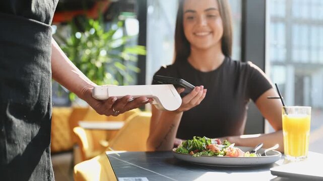 A server presents a portable card reader as a customer taps a smartphone for a contactless mobile payment while enjoying a fresh salad at a modern casual restaurant table.