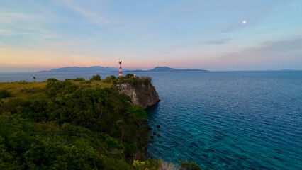 Lighthouse on a coastal cliff overlooking a calm tropical sea at sunset. The lighthouse stands above crystal clear water, creating a dramatic seascape with distant islands on the horizon. © Houston