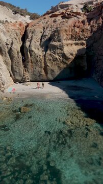 A couple discover Tsigrado Beach in Milos, Greece, where turquoise waters meet dramatic cliffs. Visitors enjoy the serenity and natural beauty of this captivating location in the Cyclades