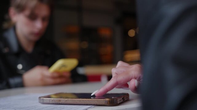 Two people interact with smartphones at wooden table in cafe setting, one gesturing toward device on surface while holding another phone, warm ambient lighting in background.