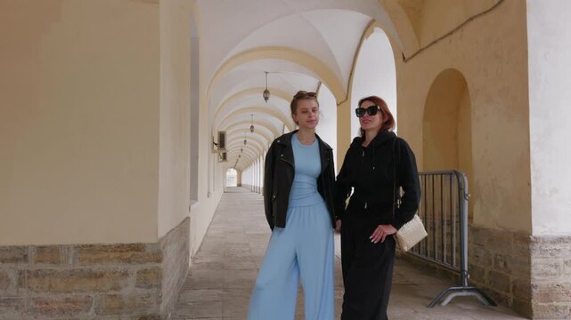 Two women stroll through elegant arched passageway with repetitive white stone columns, natural light streaming through archways in architectural setting.