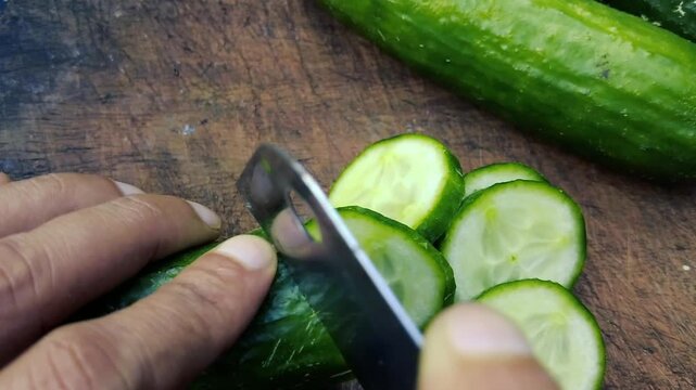 A close up of a person's hand using a knife to slice a fresh green cucumber on a wooden cutting board
