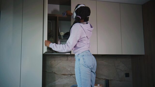 A woman reaches up to clean a kitchen cabinet while standing on a step stool. She wears headphones and focuses on her task in the kitchen.