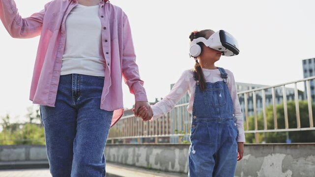 A Caucasian mother holds her daughter's hand as the child wears a virtual reality headset, experiencing a new digital world outdoors