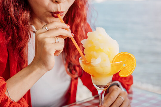 Woman with red hair drinking a refreshing frozen orange cocktail with a straw, sitting at a cafe table with a sea view